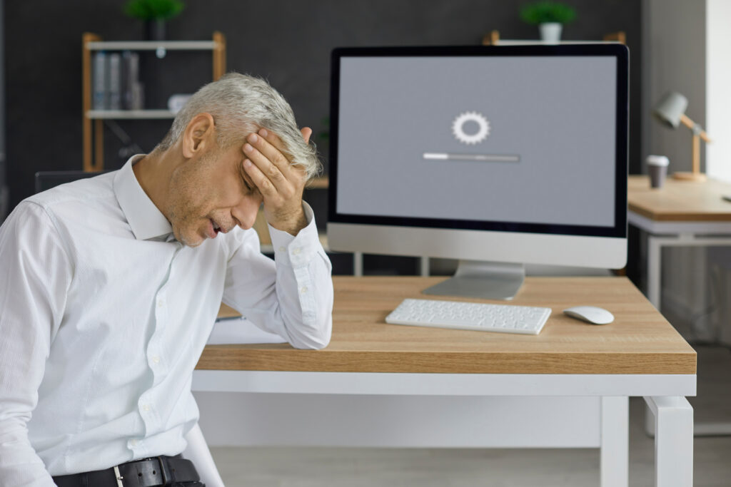 An older man sits in a chair slumped over in frustration with his hand on his forehead. A computer screen in the background shows a website that's loading too slowly. 