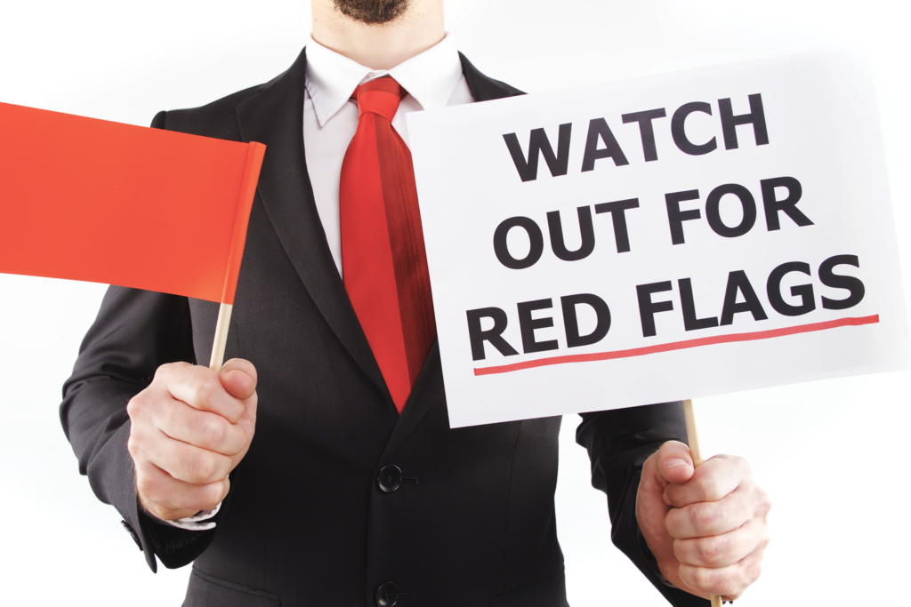A man in a black suit with a two-tone red tie holds a red flag in his right hand and a sign in his left hand that says "Watch Out For Red Flags."