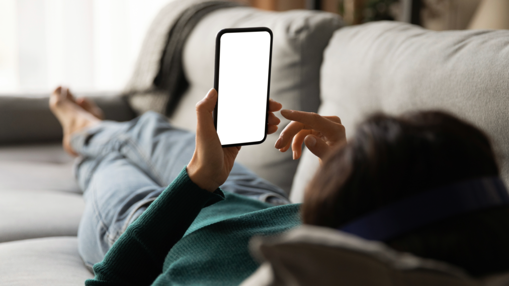 A woman lays on a couch and uses her mobile phone to access the internet. 